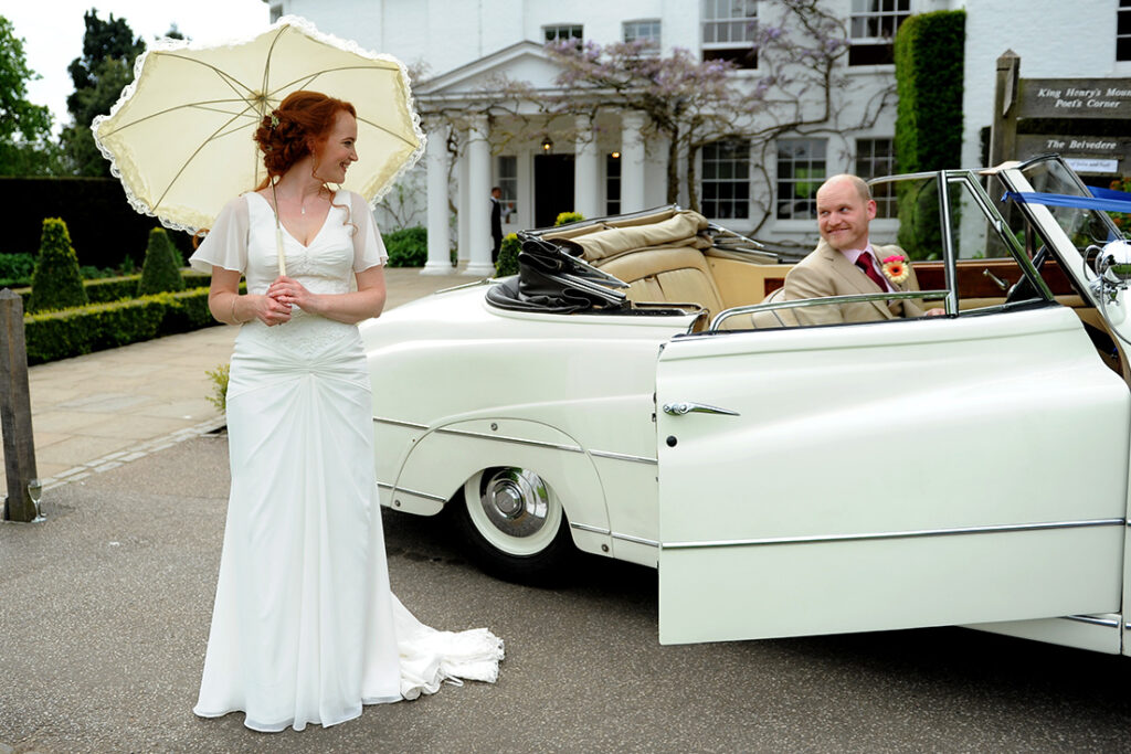 A bride holding a lace parasol looking back at a man in a vintage car.