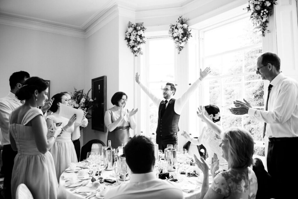 A black and white photo of a wedding party, featuring the bride, groom, and guests in formal attire, smiling together.