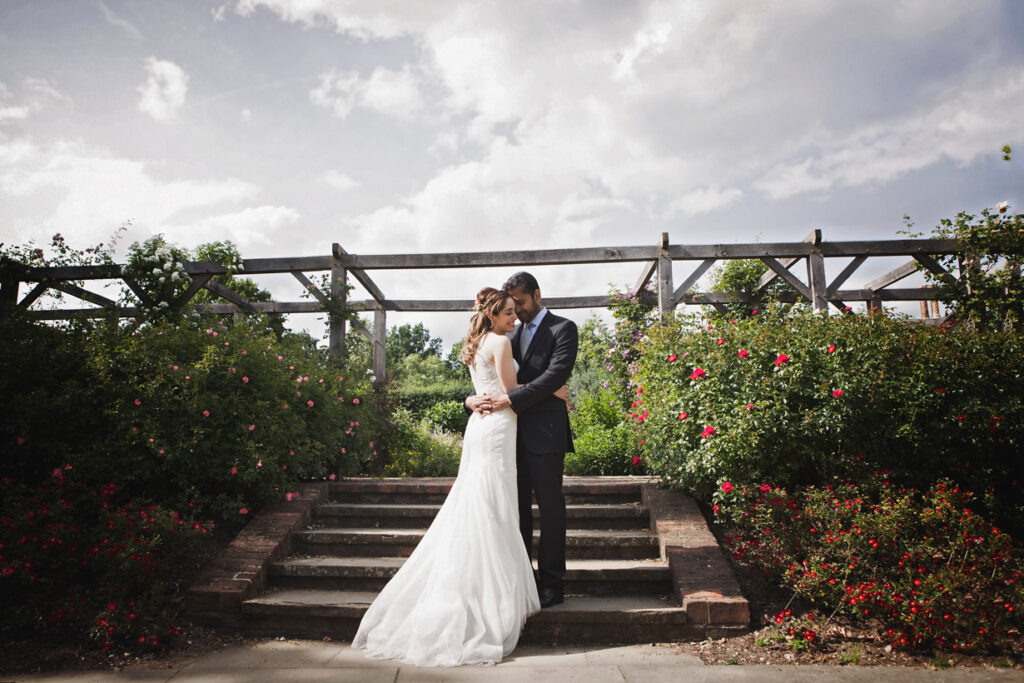 A bride and groom embrace on stone steps surrounded by rosebushes.