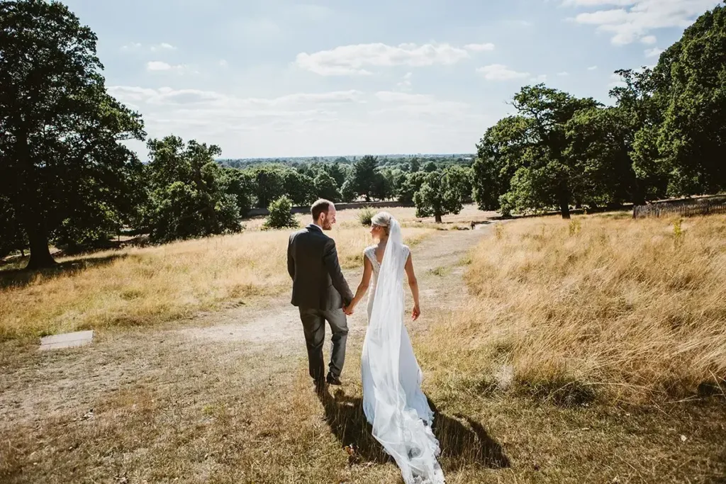 A bride and groom holding hands and walking through parkland on a sunny day.
