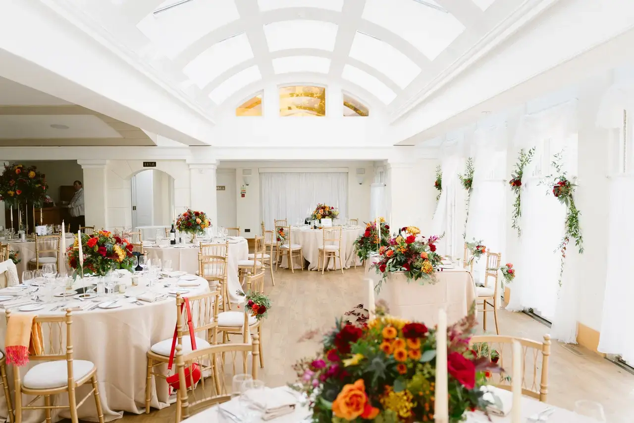 A large brightly lit room with tables, chairs and floral arrangements.