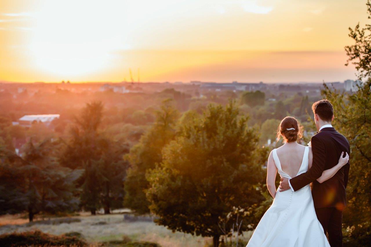 A bride and groom looking down onto trees and fields at sunset.