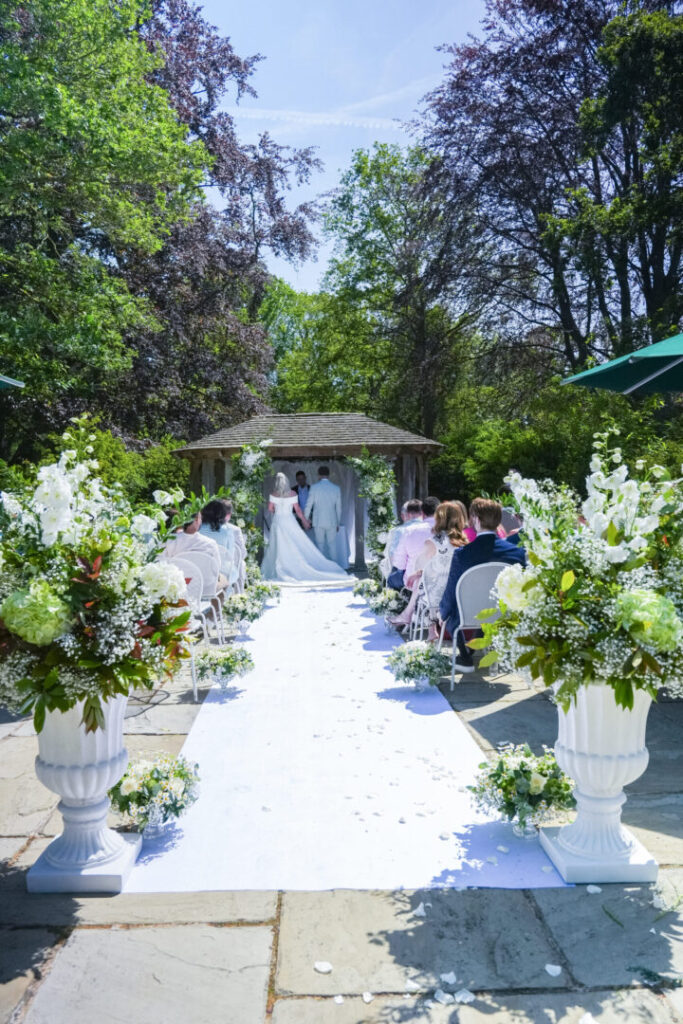 Outdoor wedding ceremony in a garden, featuring an arch adorned with flowers and guests seated on white chairs.
