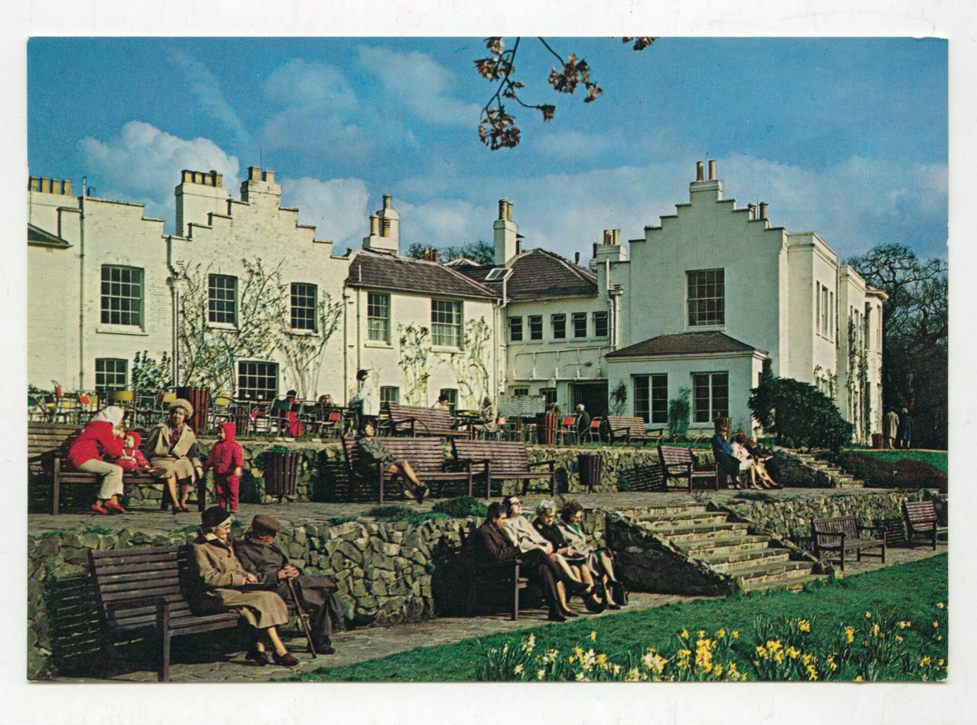 Vintage 1970s postcard depicting Pembroke Lodge in Richmond Park, with people seated on the landscaped terraces and gardens outside the historic white Georgian house under a blue sky.