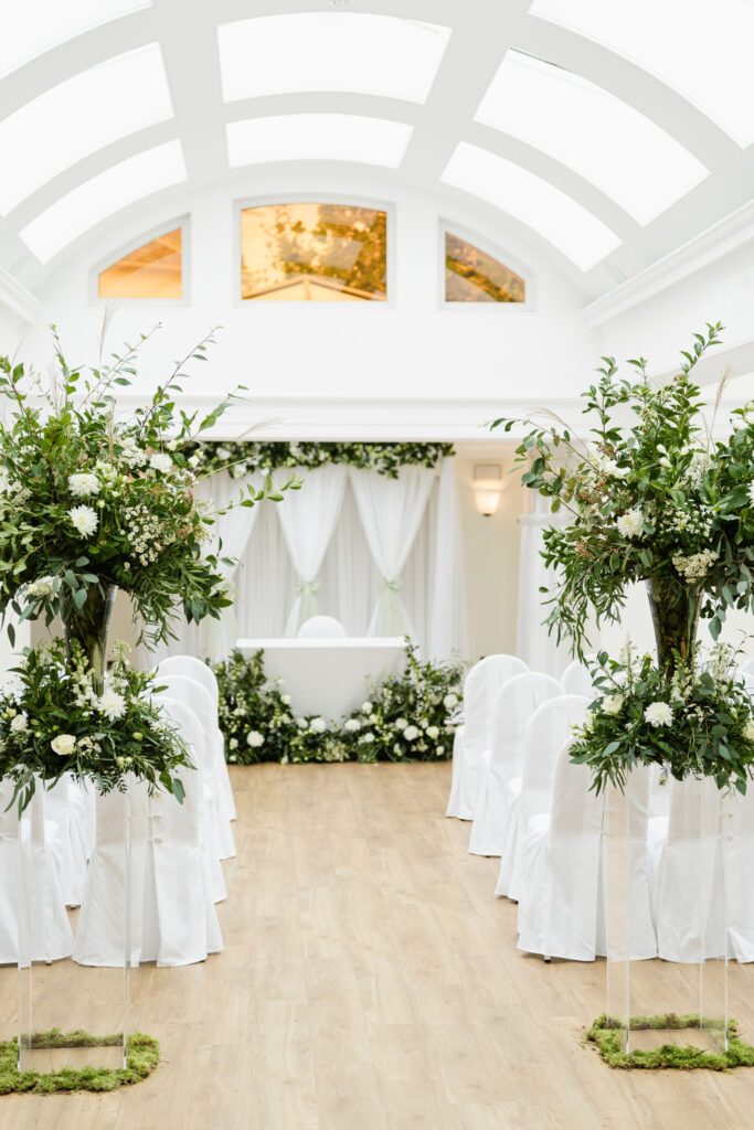 A brightly lit room with a large glass ceiling with rows of chairs and floral arrangements.